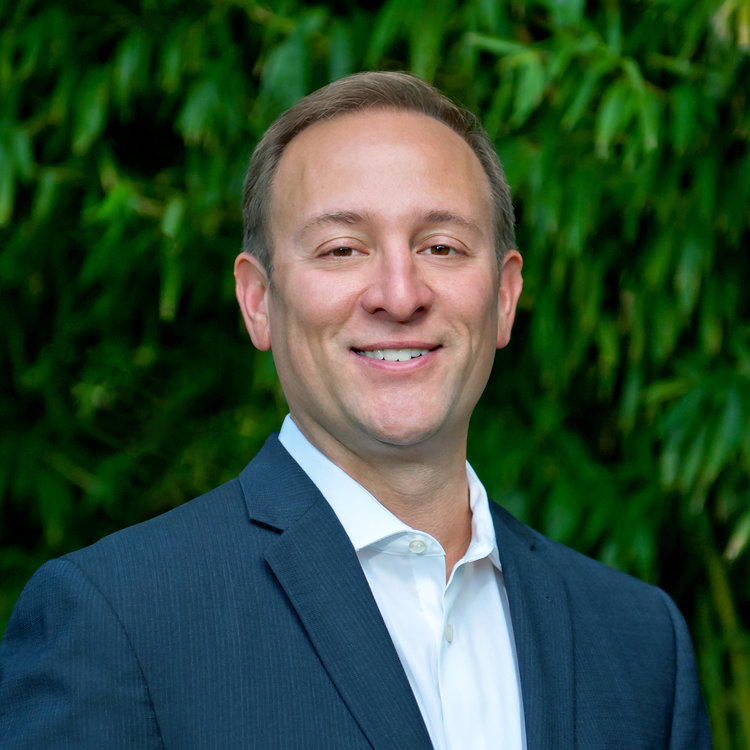 A strategic man in a suit smiles in front of bushes.