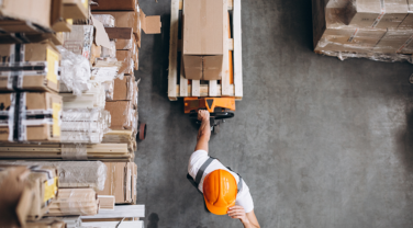 A company worker in an orange hard hat is strategically operating a forklift in a warehouse.