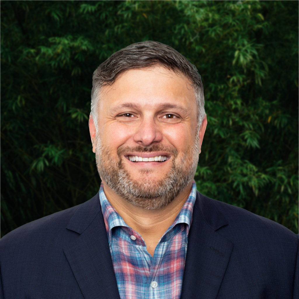 A man with a strategic smile standing in front of a tree.